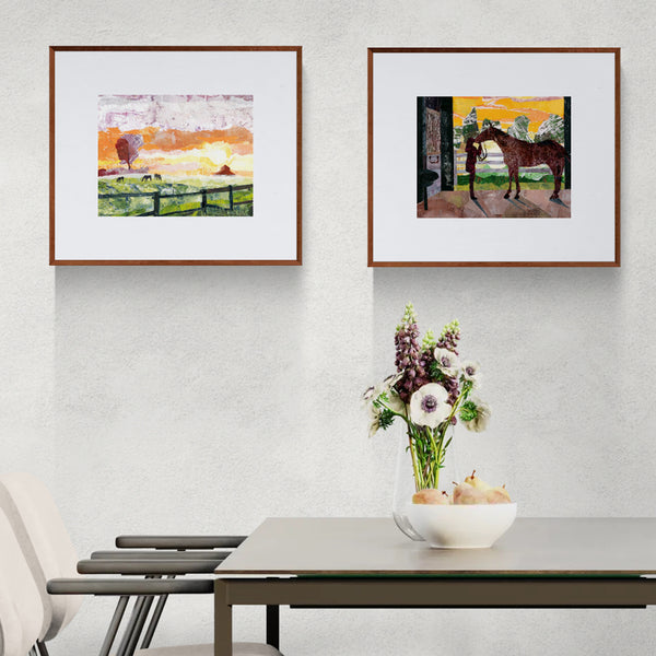 Two framed art prints hang on a light gray textured wall above a dining table with a vase of flowers. The left print shows a colorful sunrise landscape with horses grazing, and a barn in the distance, and the right print shows a horse standing in a doorway of a barn with a woman at sunrise.